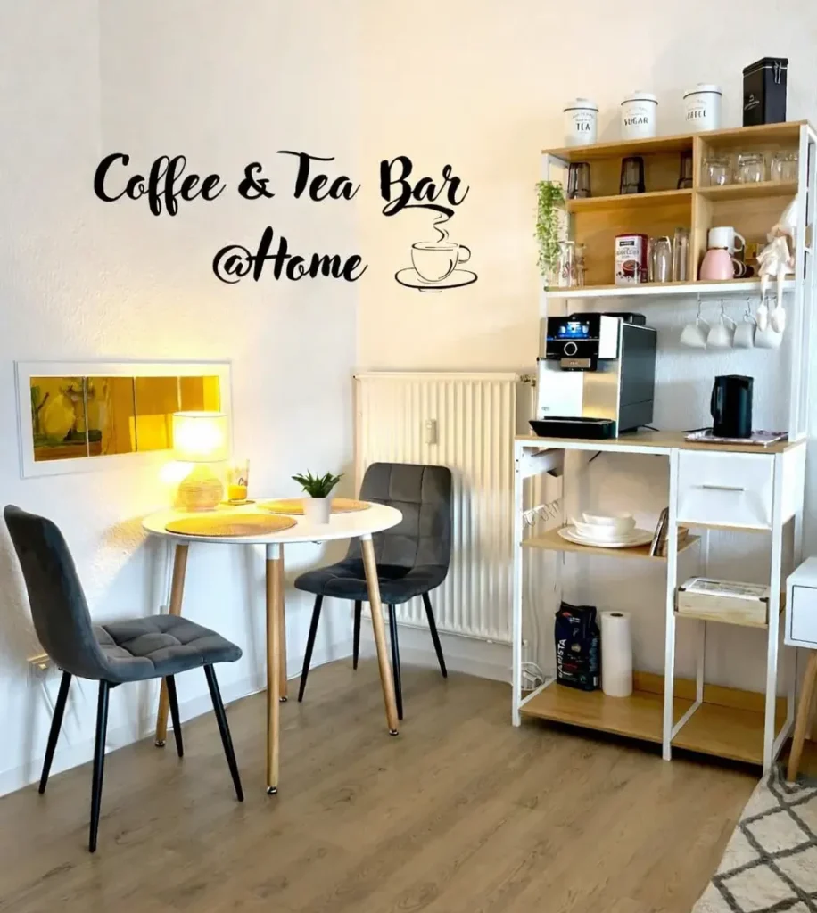 Helle Kaffee-Bar im Wohnraum mit rundem weißen Tisch, zwei grauen Stühlen, weißem Kaffee-Regal aus Holz und Metall, Kaffeemaschine, Tassen und Wandbeschriftung „Coffee & Tea Bar @Home“.